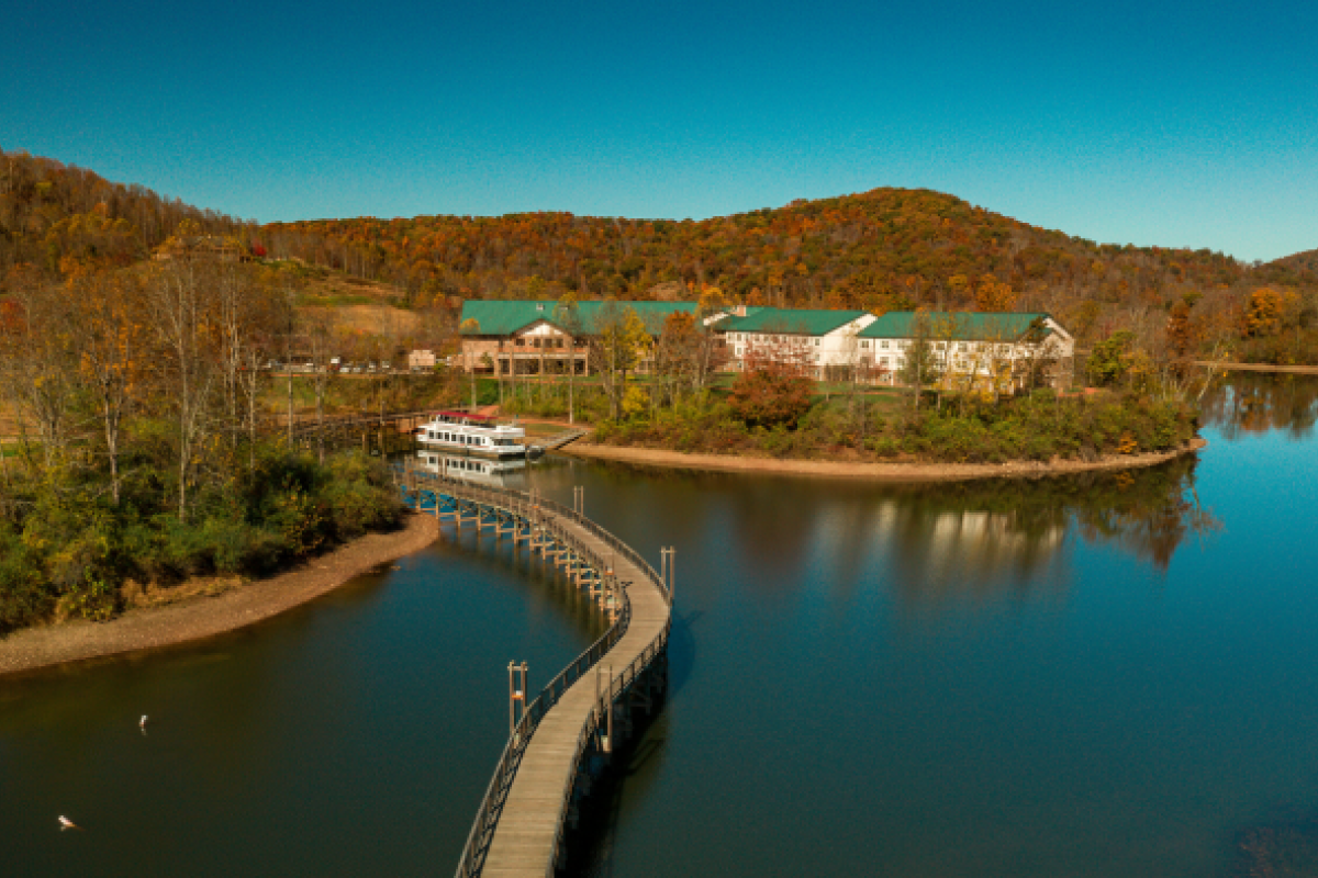 This image shows a scenic view of a lake with a curved walkway leading to buildings surrounded by trees and hills under a clear blue sky.