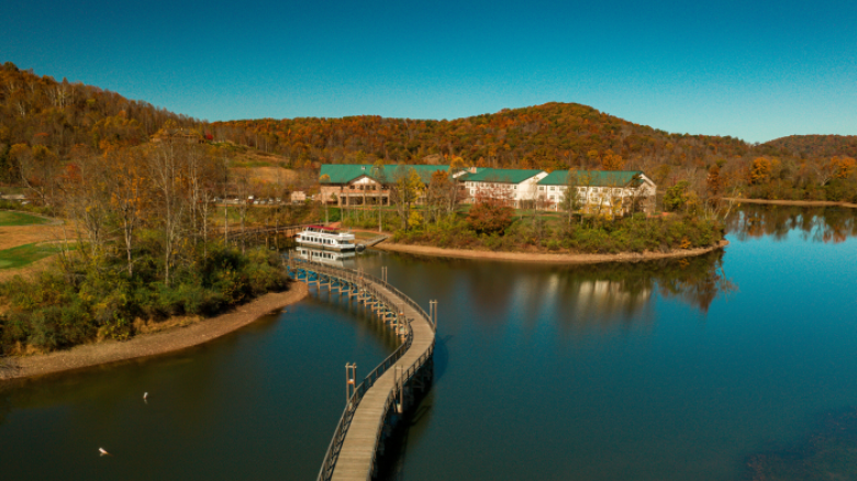 This image shows a scenic view of a lake with a curved walkway leading to buildings surrounded by trees and hills under a clear blue sky.