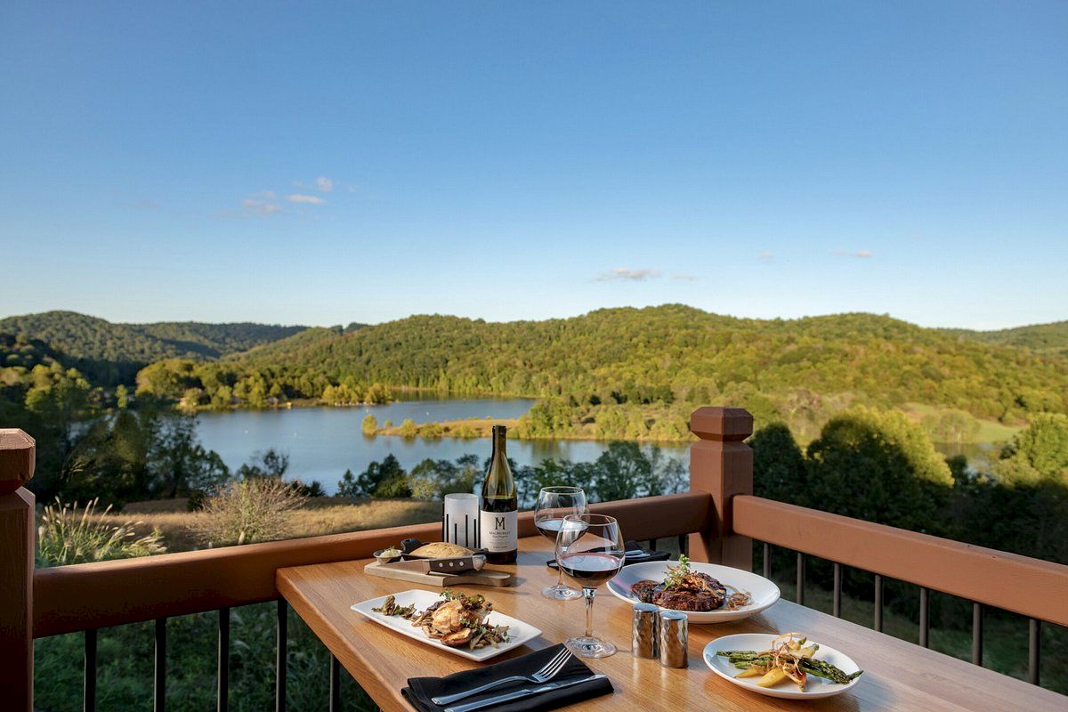 A scenic outdoor dining setup on a wooden balcony, showcasing a beautiful lake and green hills, with plates of food, wine, and glasses on the table.
